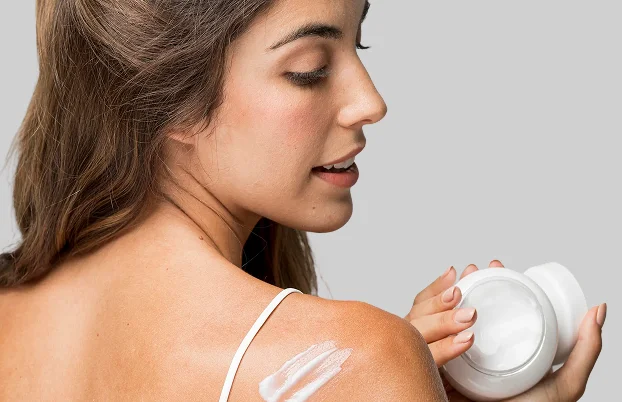 Woman applying moisturizing cream to her shoulder while holding skincare jar